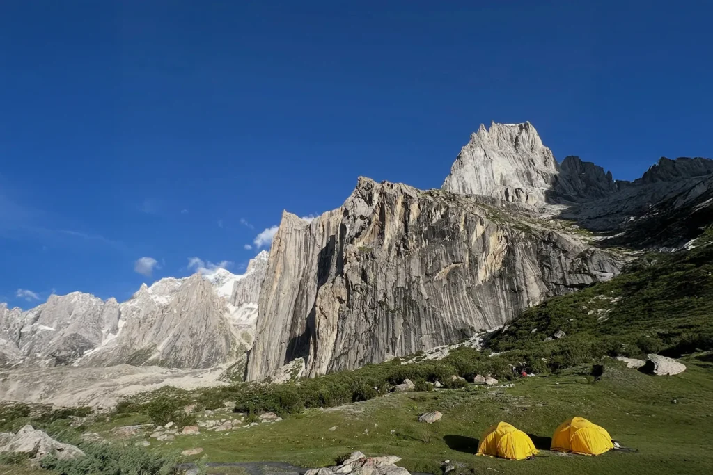 Trekkers camp in a green meadow below granite towers with a glacial stream, on the Charakusa Valley trek